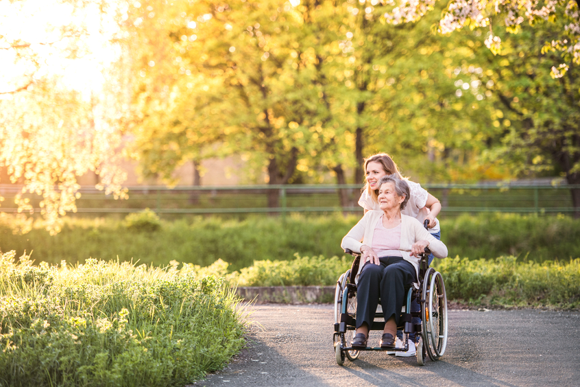 Jüngere Frau schiebt ältere Frau im Rollstuhl durch einen Park/Garten