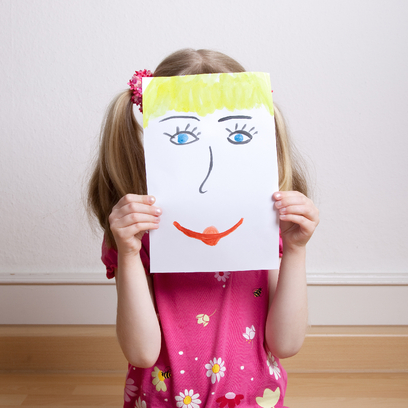 Mädchen mit Papier vor dem Gesicht; auf Papier ist mit Buntstiften Gesicht einer Frau/Mädchen gezeichnet