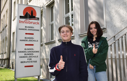 Lia und Tobias vor dem Eingang der Handelsakademie, mit Daumen nach oben.
